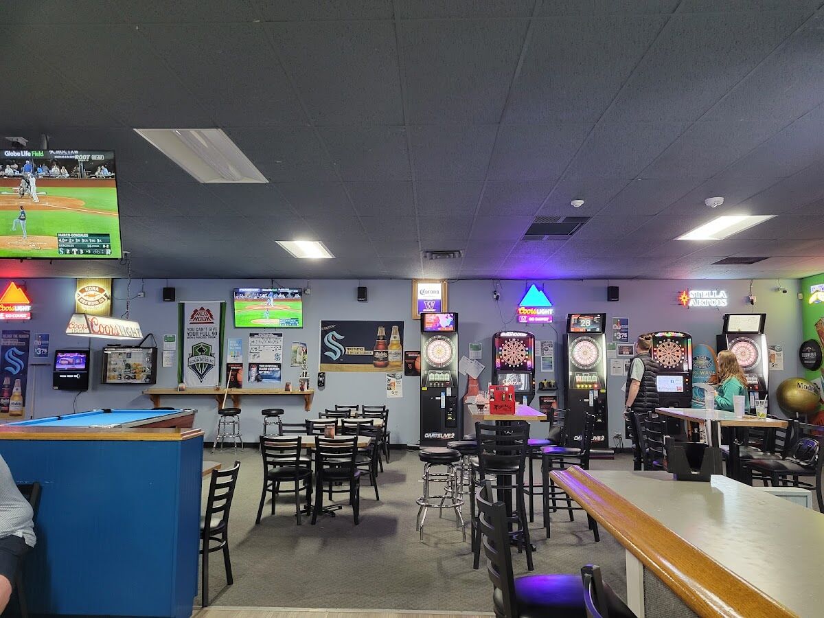Inside Just Chill Pub & Grill: a pool table, three DartsLive electronic dart machines, big screens showing baseball and Seahawks, Sounders and Kraken banners on the wall.