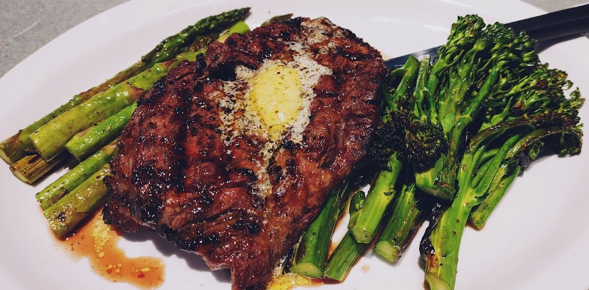 A 12 oz ribeye at Just Chill, char-marked with a melting herb butter, plated with grilled asparagus and broccolini.